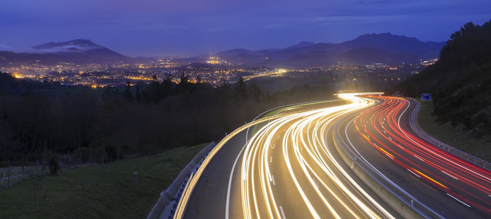 Highway at night with white and red blurred lines representing fast-moving cars with mountains in background