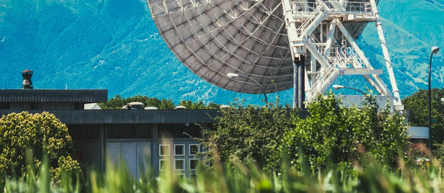 Large satellite in a lush green field faces mountains on a bright blue day. Positioned next to a single small house.